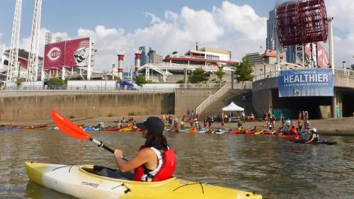A person kayaking on a river with a city skyline and a gathering of kayakers in the background.