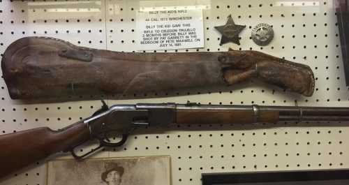 A display featuring a leather gun holster, a Winchester rifle, and vintage badges on a pegboard background.