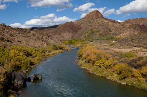 A serene river flows through rocky terrain, surrounded by colorful autumn foliage and under a blue sky with clouds.