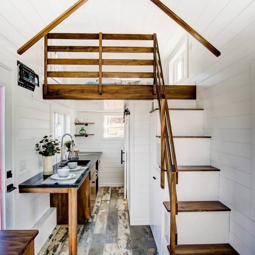 A cozy tiny house interior featuring a lofted bed, wooden stairs, and a modern kitchen area with natural light.