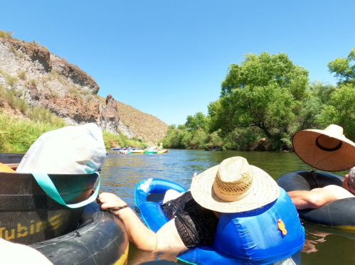 People relaxing on inner tubes in a river, surrounded by lush greenery and rocky cliffs under a clear blue sky.
