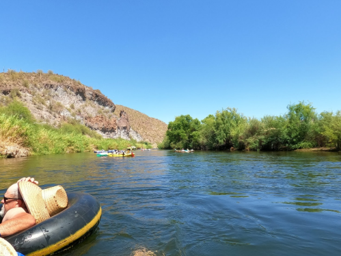 A person relaxes on an inner tube in a river, surrounded by lush greenery and rocky hills under a clear blue sky.