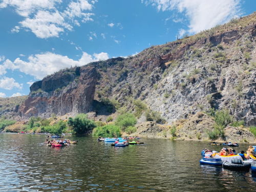 A group of people in colorful rafts floating on a river, surrounded by rocky cliffs and greenery under a blue sky.