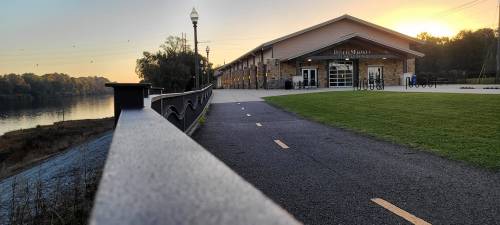 A scenic view of a pathway by a river, leading to a modern building at sunrise.