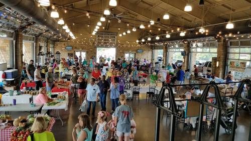 A bustling indoor market filled with people browsing stalls, enjoying food, and socializing under string lights.