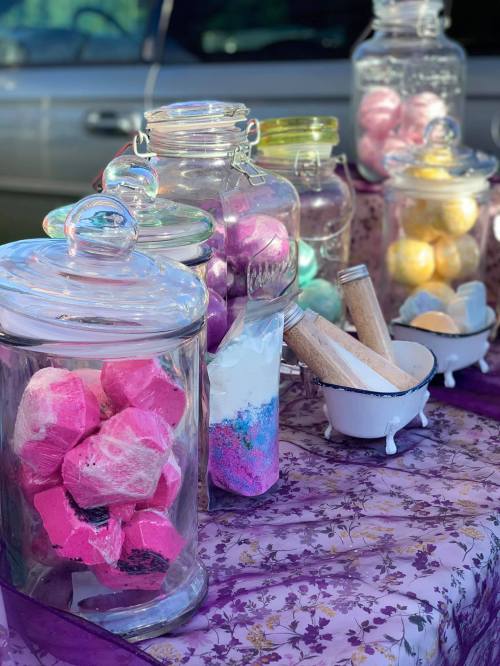 Colorful bath bombs and salts displayed in glass jars on a purple floral tablecloth.