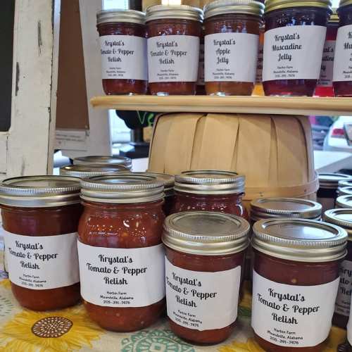 Jars of various relishes and jellies labeled "Krystal's Tomato & Pepper Relish" displayed on a wooden shelf.