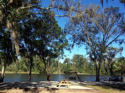 A serene riverside scene with picnic tables, trees, and clear blue skies reflecting on the water.