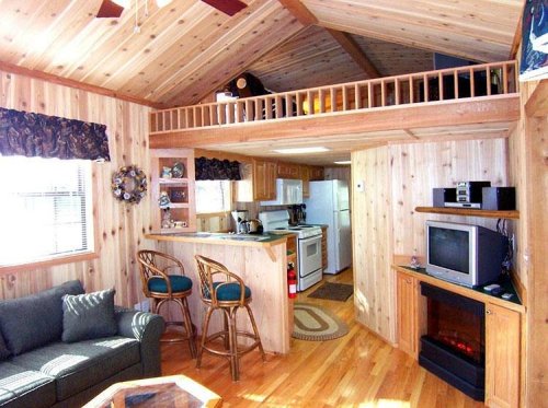 Cozy wooden cabin interior with a living area, kitchen, and loft, featuring warm wood tones and comfortable furnishings.