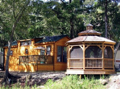 A cozy wooden cabin next to a gazebo, surrounded by trees and greenery.