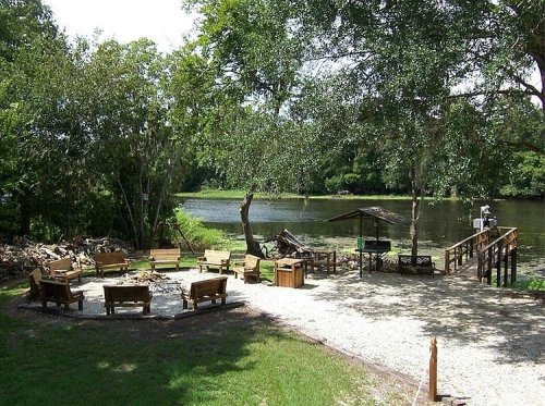 A serene riverside area with wooden benches arranged in a circle, surrounded by trees and a small dock.