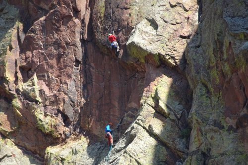 Two climbers ascend a rocky cliff, one near the top in red and the other lower down in blue.
