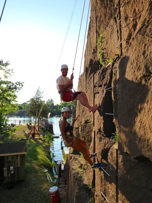 Two climbers ascend a rock wall by a river, surrounded by trees and a clear blue sky.