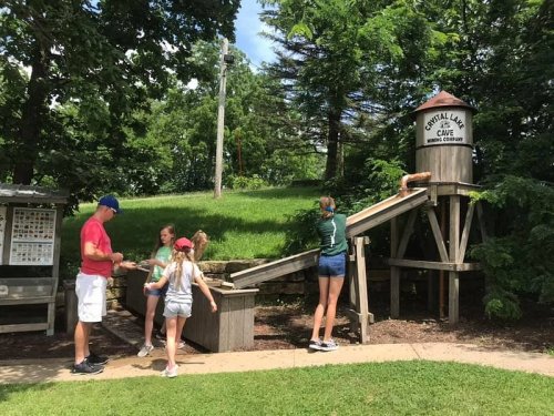 Children play on a wooden slide near a water tower structure at a park, with adults supervising nearby.