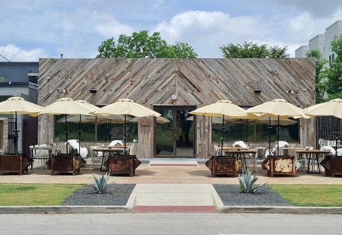 A rustic restaurant with wooden exterior, outdoor seating under yellow umbrellas, and green landscaping.