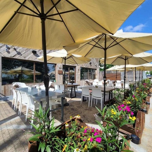 Outdoor dining area with white chairs, large yellow umbrellas, and colorful flowers in planters. Bright, sunny atmosphere.