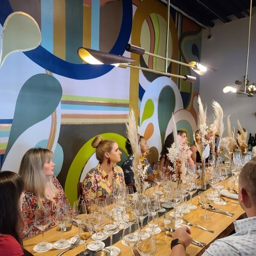A long dining table set with glasses and decorative plants, with guests seated against a colorful mural backdrop.