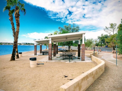 A sandy area by a lake featuring picnic shelters, palm trees, and a clear blue sky.