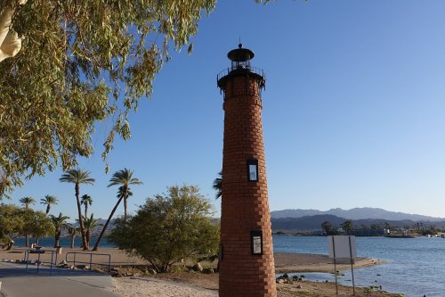A brick lighthouse stands by a calm waterway, surrounded by palm trees and mountains in the background.