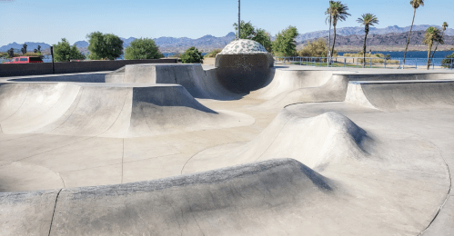 A concrete skatepark with smooth curves and a dome feature, surrounded by palm trees and mountains in the background.