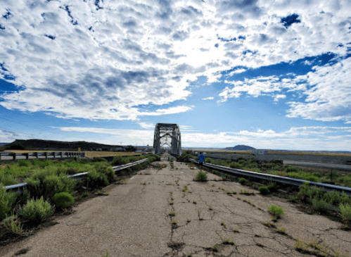 An abandoned bridge stretches into the distance under a cloudy sky, surrounded by overgrown vegetation.
