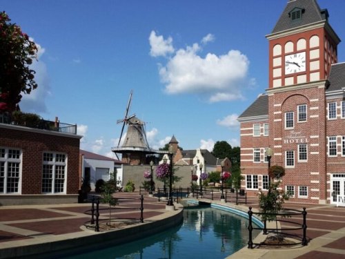 A charming hotel with a clock tower beside a canal, featuring a windmill and blooming flowers under a blue sky.