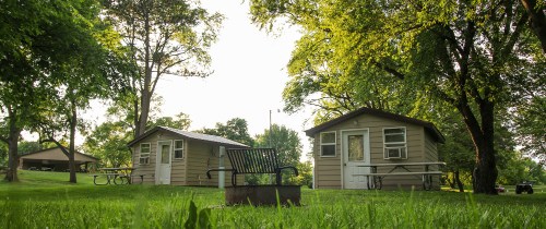 Two small cabins surrounded by trees and grass, with a bench and picnic tables nearby in a serene outdoor setting.
