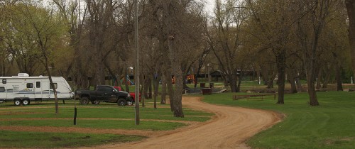A dirt road winds through a park with trees, an RV, and a truck parked nearby.