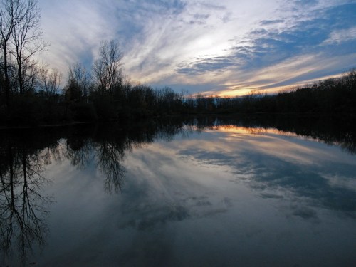 A serene lake at sunset, reflecting colorful clouds and trees against a tranquil water surface.