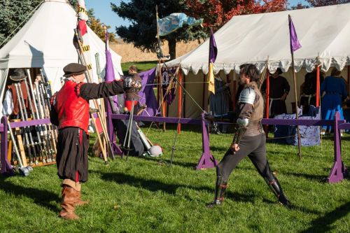 Two men in medieval costumes spar with swords in a grassy area, surrounded by tents and colorful banners.