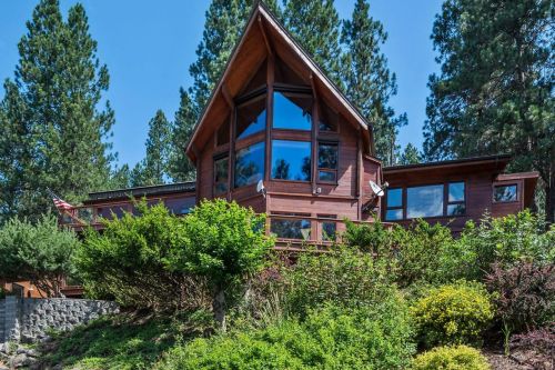 A modern wooden house with large windows, surrounded by greenery and tall trees under a clear blue sky.