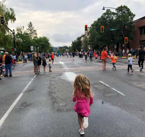 A young girl in a pink dress runs down a wet street during a lively community event with many people in the background.