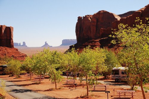 A scenic view of red rock formations and green trees in a desert landscape, with benches and an RV park in the foreground.