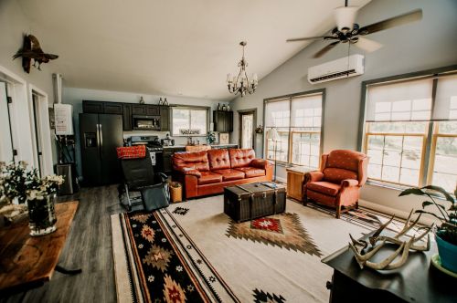 Cozy living room with a red sofa, armchair, wooden coffee table, and kitchen area, featuring large windows and rustic decor.