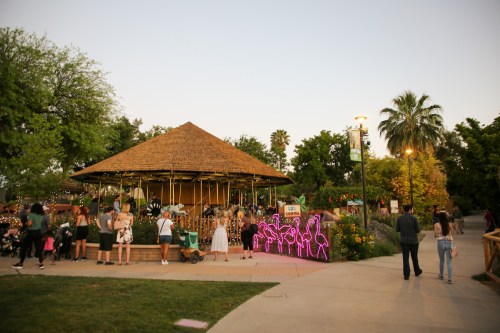 A lively park scene with a thatched-roof gazebo, colorful lights, and people enjoying the evening.