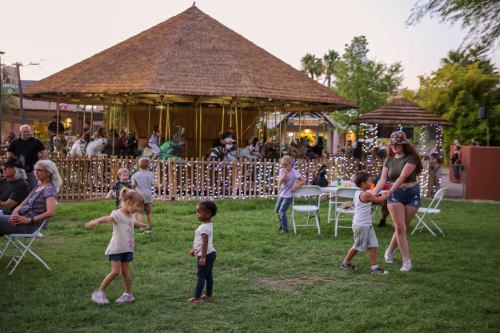 Children play and dance on a grassy area near a thatched-roof pavilion, with people seated and festive lights in the background.