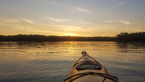 A kayak on calm water at sunset, with golden hues reflecting on the surface and trees in the background.