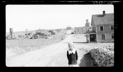 A young girl stands on a dirt road, with a small town and buildings in the background under a clear sky.
