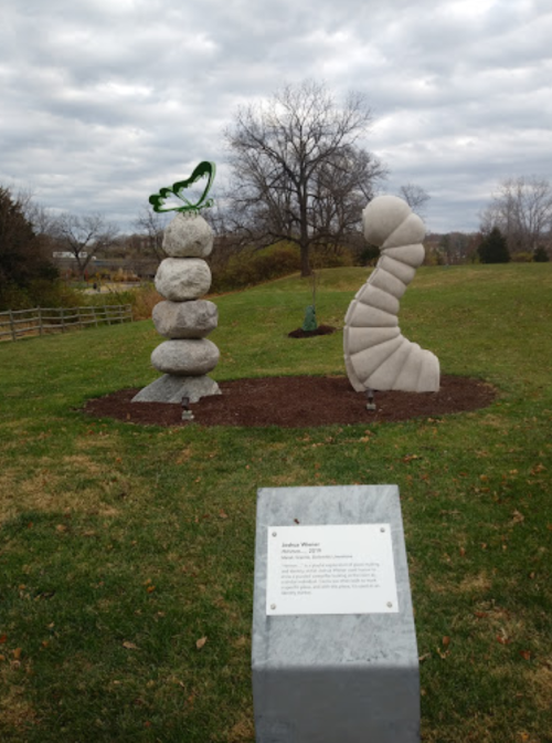 Two abstract sculptures in a grassy area: one resembles stacked stones with a green butterfly, the other a spiral form.