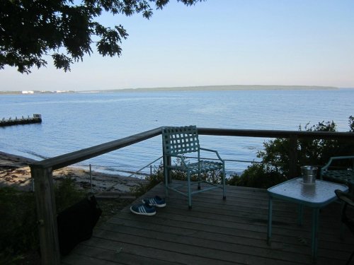 A peaceful lakeside view from a wooden deck with chairs, a table, and a distant shoreline under a clear sky.