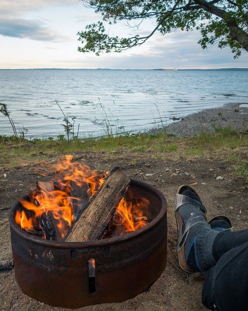 A cozy campfire burns in a metal ring by a serene lakeside, with a person’s feet resting nearby on the sandy shore.