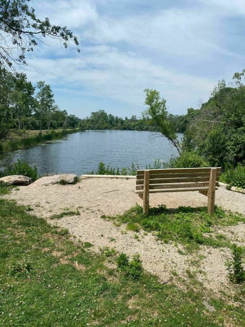 A wooden bench on a gravel area overlooking a calm lake surrounded by trees under a partly cloudy sky.