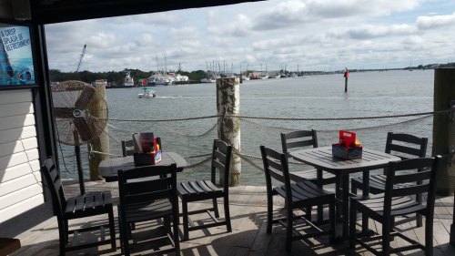 Outdoor dining area with black tables overlooking a calm waterway and boats in the distance under a cloudy sky.