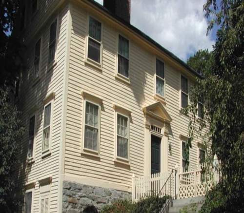 A historic two-story house with a light-colored exterior, multiple windows, and a front staircase surrounded by greenery.