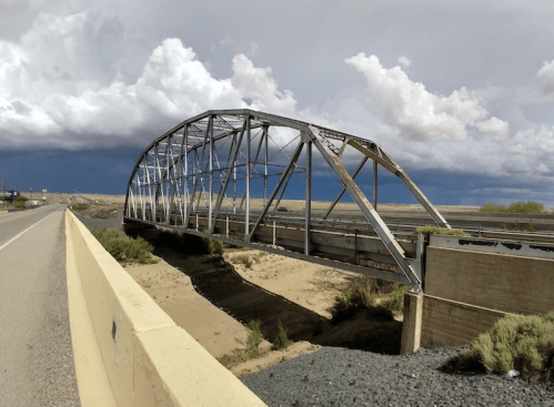 A weathered metal bridge arches over a dry riverbed, with dark storm clouds looming in the background.