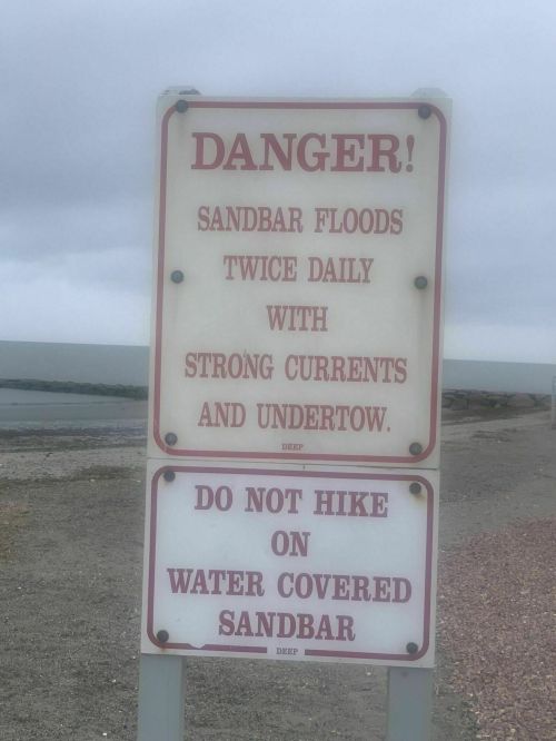 Sign warning of danger due to sandbar flooding, strong currents, and advising against hiking on water-covered sandbars.