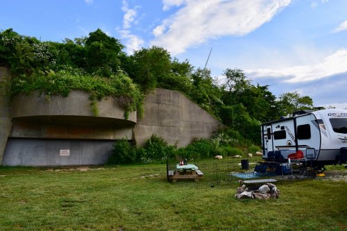 An RV parked near a large, overgrown concrete structure, surrounded by grass and scattered rocks under a cloudy sky.