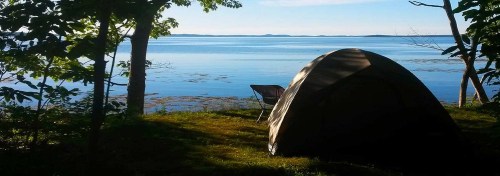 A tent by a calm lake, surrounded by trees, with a clear blue sky and distant hills in the background.