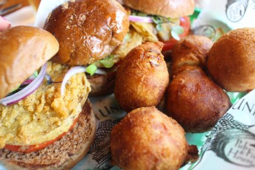 Two burgers with toppings and a side of golden, round fritters on a patterned paper.