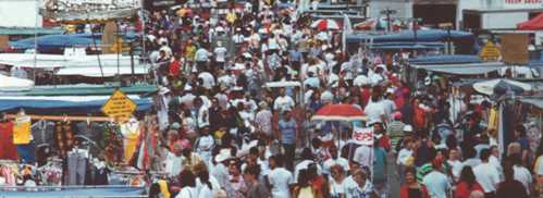 A bustling market scene filled with crowds of people, colorful tents, and various stalls under a bright sky.